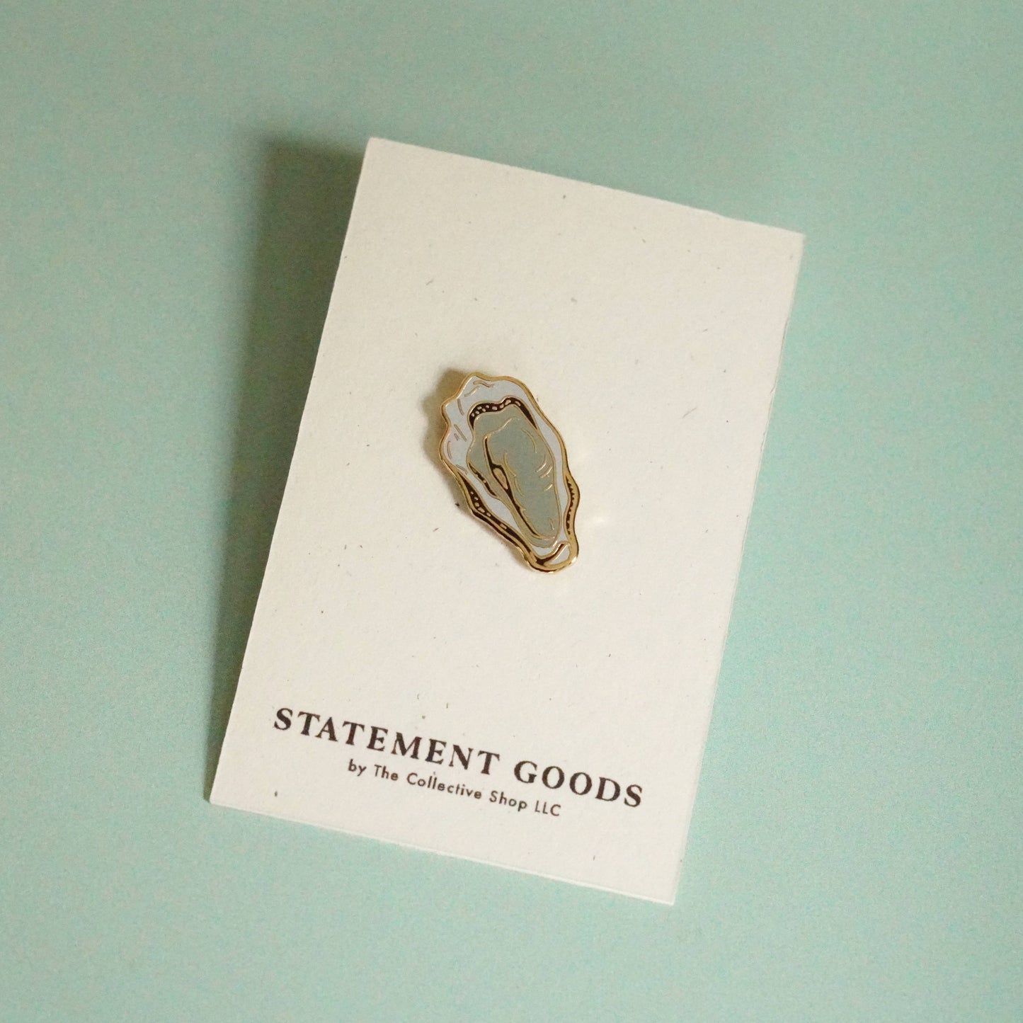 Calling all oyster lovers! This playful pin features a gold plated, hard enamel oyster illustration by local artist Statement Goods. Its compact size of 1-1/8" x 5/8" makes it the perfect addition to any pin collection. Secured with a black rubber backing.