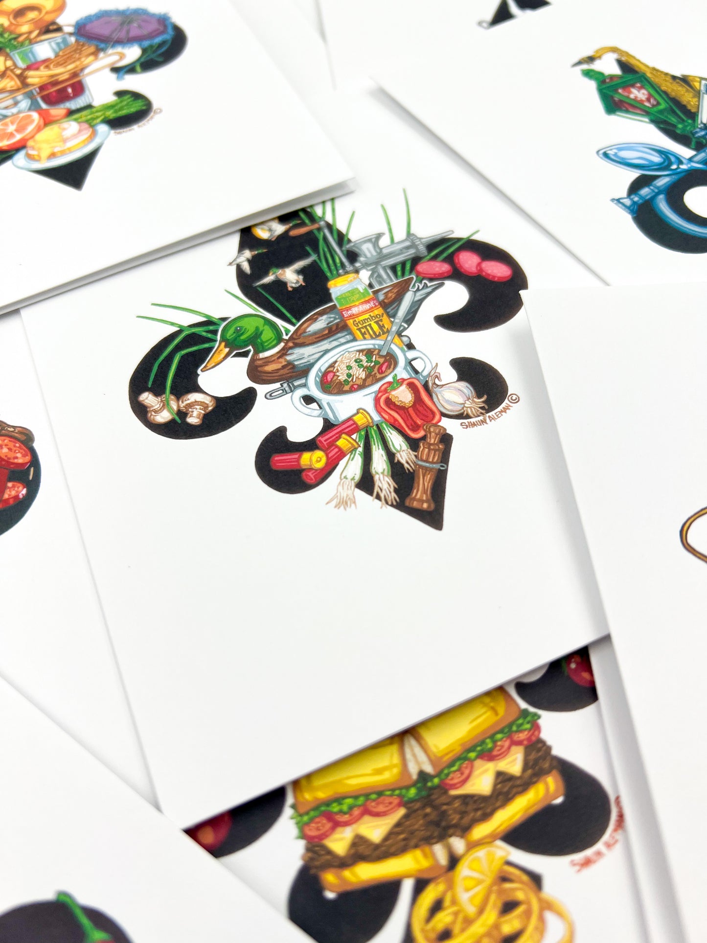 A close-up of several white cards or envelopes scattered on a surface, each printed with a colorful illustrated food graphic. The central card shows a stylized chef/cooking illustration with vegetables, utensils, and a bowl, while other cards show partial food designs like a sandwich and pasta.
