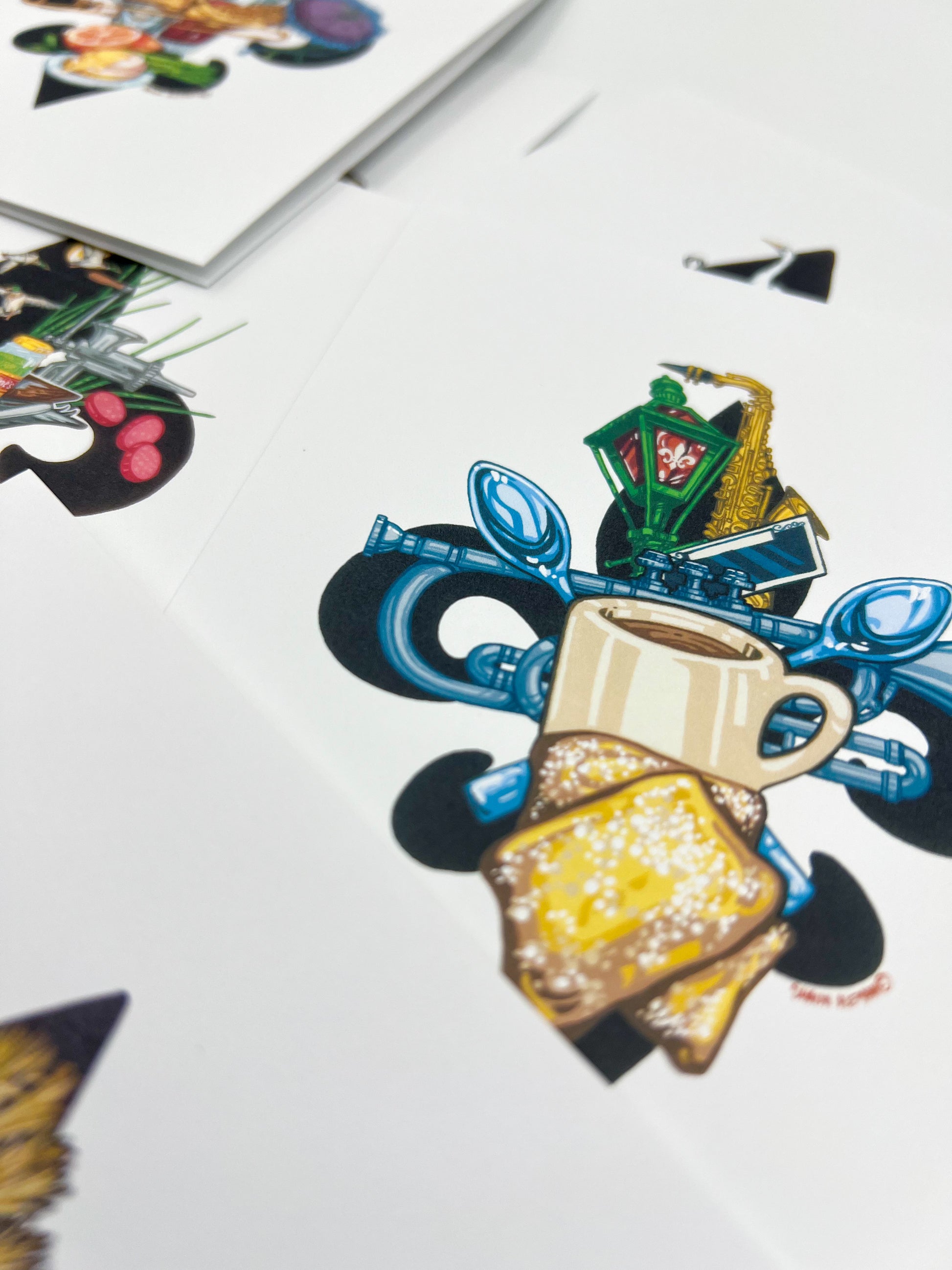 A close-up of printed artwork or packaging featuring a stylized illustration of a beige coffee mug, beignets, green lantern, and blue spoons and trumpet elements on a white surface.